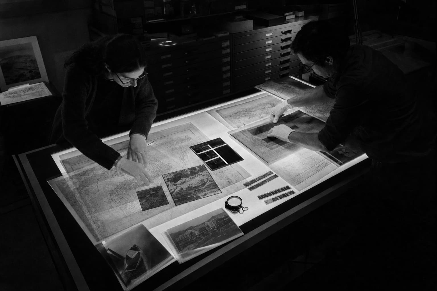 Two archivists arrange maps and photographic records across a glowing table in a dark collection room